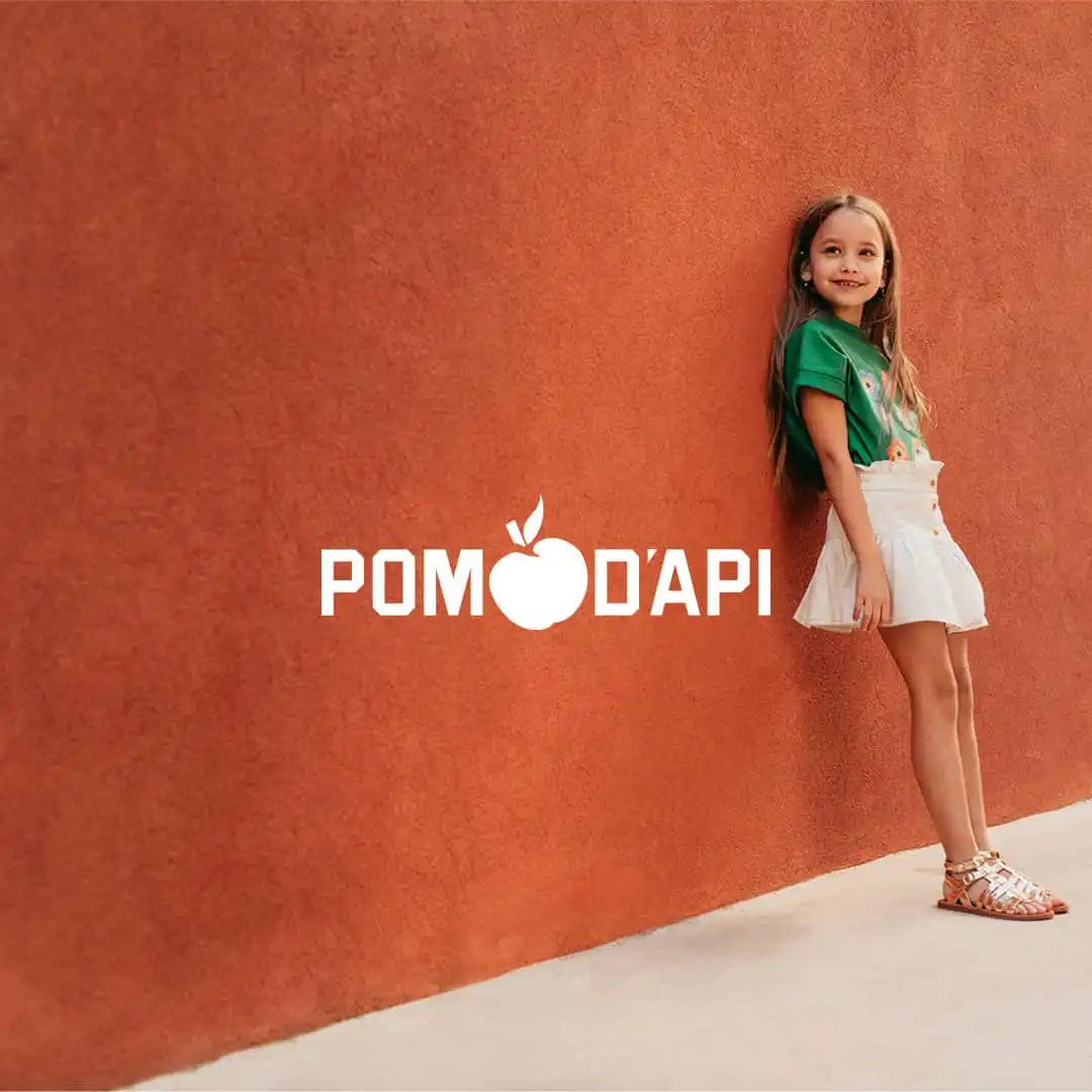 Un enfant portant une chemise verte et une jupe blanche se tenant contre un mur en terre cuite avec le texte 'Pomodapi'.