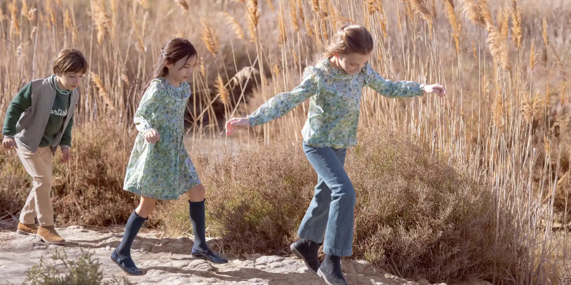 Three children walking in a line along a narrow dirt path wearing casual clothes.