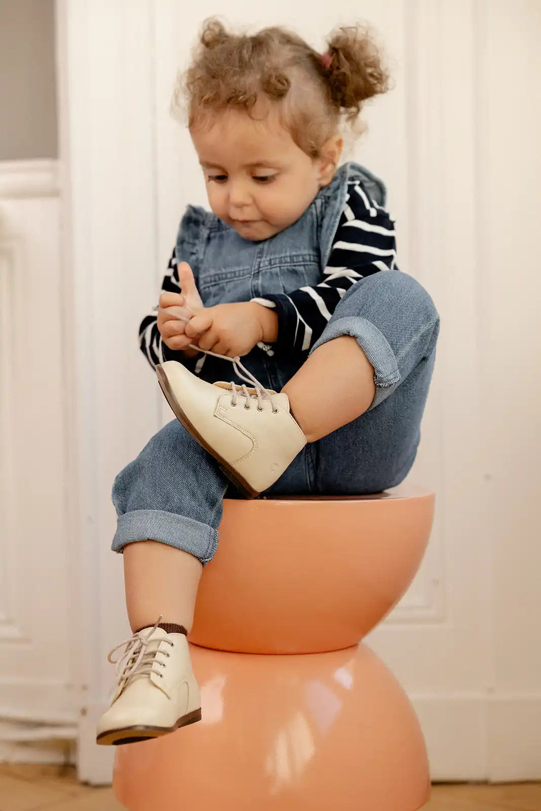 Un enfant en salopette en jean assis sur un tabouret de couleur pêche.