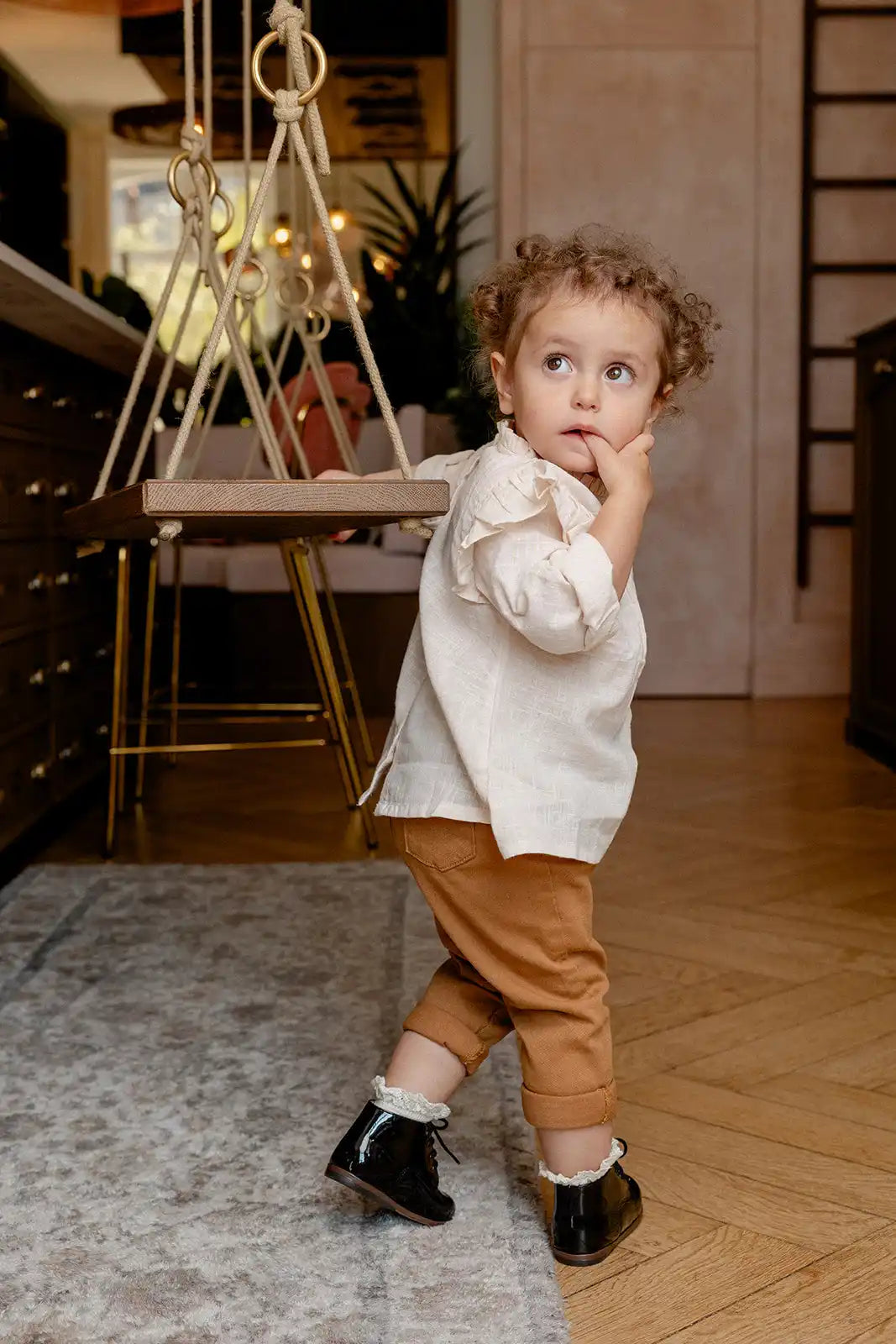 Un enfant en bas âge portant une chemise blanche et un pantalon marron se tient avec un doigt près de sa bouche.